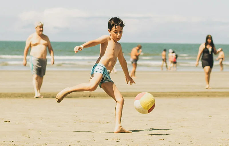 @lang('Menino a brincar com uma bola na praia, rodeado por banhistas num dia de verão, simbolizando liberdade e proteção ao ar livre')